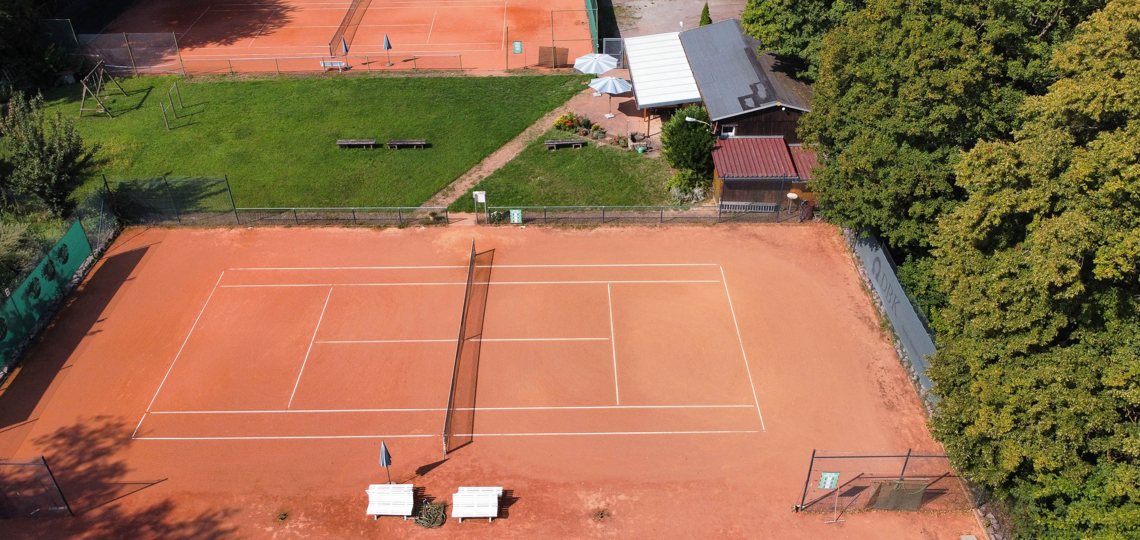 Luftaufnahme der Tennisanlage des Tennisverein Mörzheim: Ein roter Sandtennisplatz im Vordergrund, dahinter eine Grünfläche mit Bänken und das Vereinsheim mit Terrasse. Weitere Tennisplätze sind im Hintergrund sichtbar.